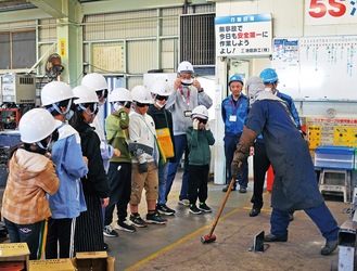 溶接した鉄の部品をハンマーで叩くことで強度を伝える社員と見学する児童たち