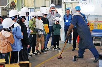 溶接した鉄の部品をハンマーで叩くことで強度を伝える社員と見学する児童たち